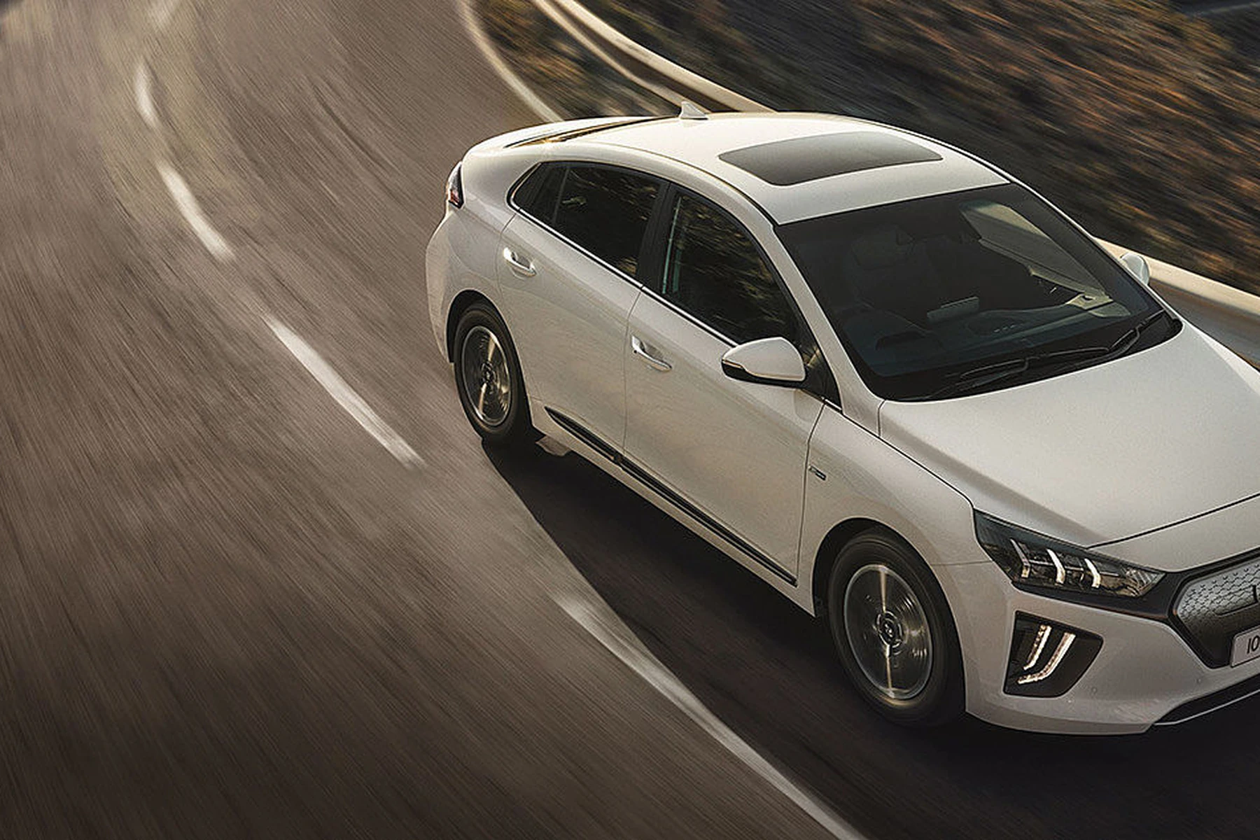 A white car driving on a winding mountain road, surrounded by rocky terrain and a clear sky.