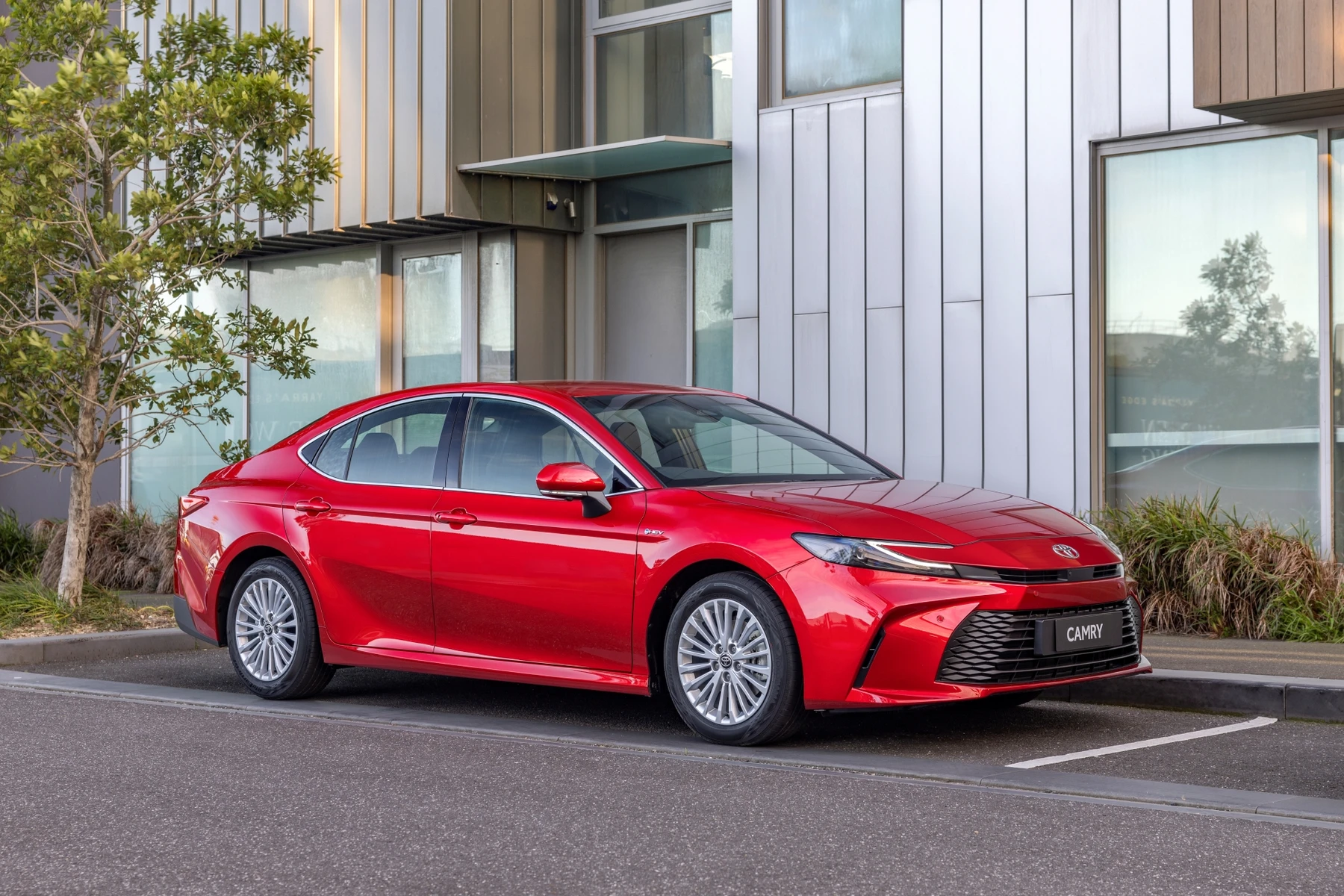 A sleek red sedan parked on the street in front of a modern building, with trees and glass windows in the background.