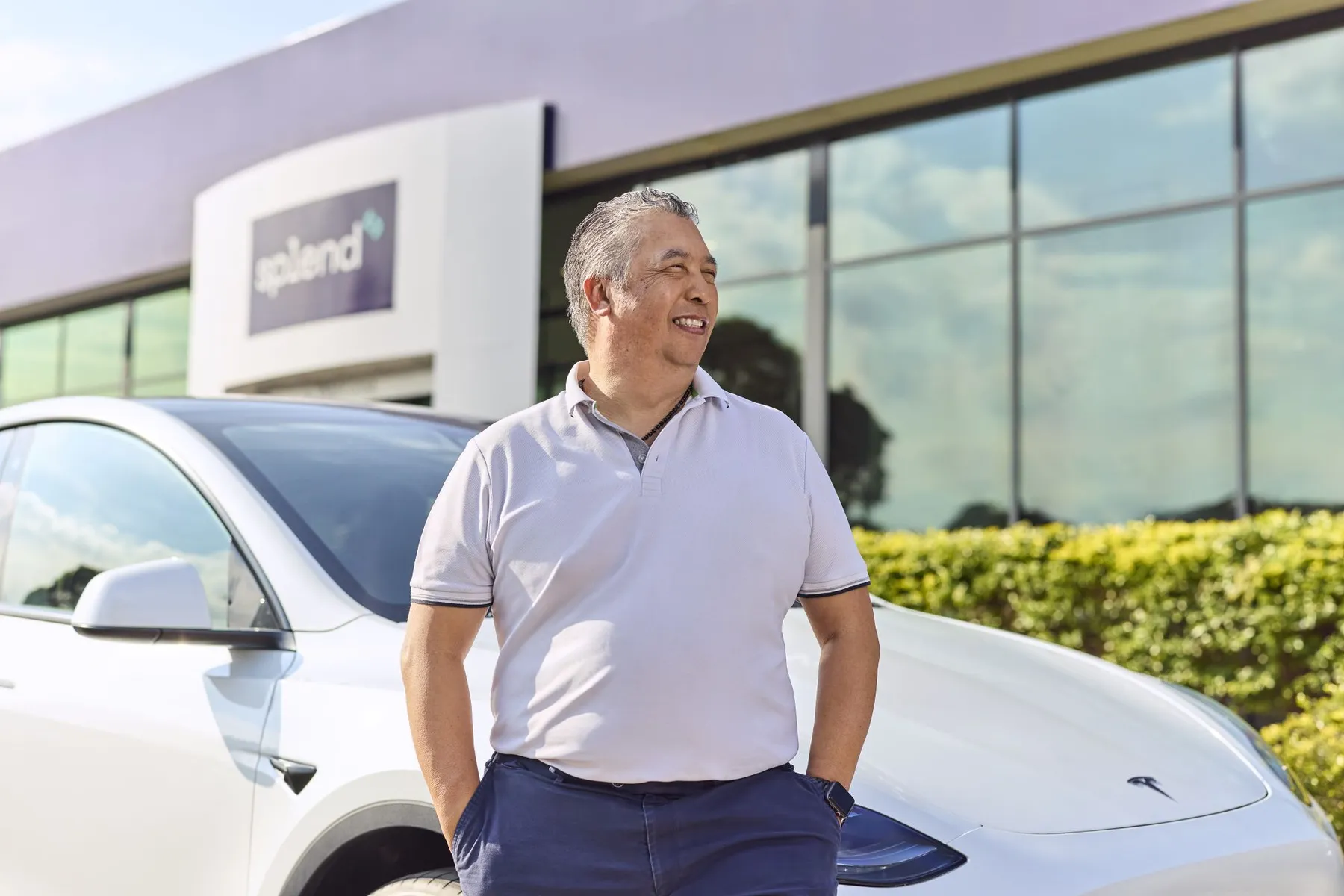 Man in a light polo shirt stands confidently beside a white Tesla, with a modern building and greenery in the background.