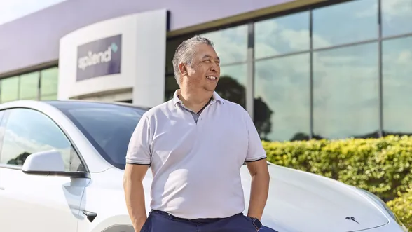 Man in a light polo shirt stands confidently beside a white Tesla, with a modern building and greenery in the background.