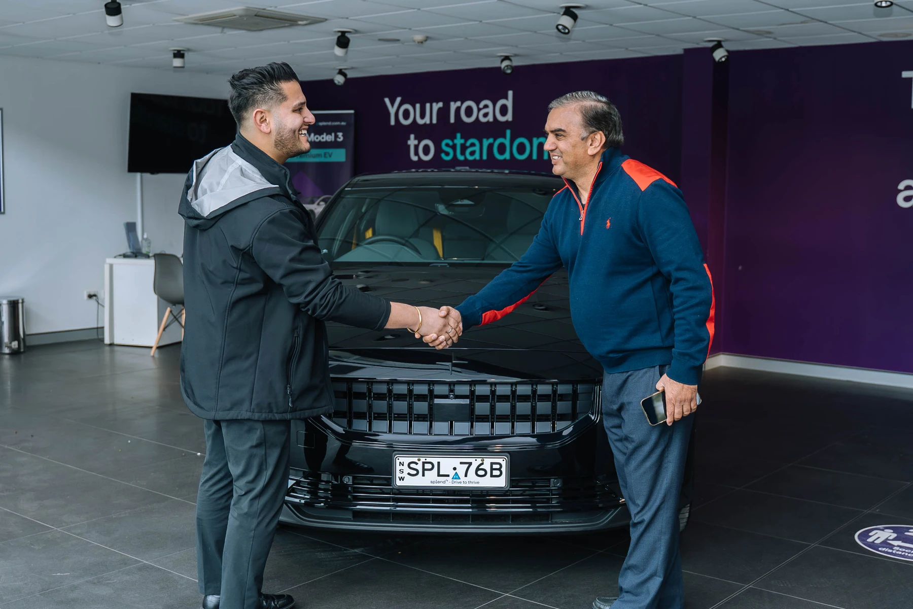 Two men shaking hands in a showroom, standing in front of a black Polestar with a visible license plate. Purple wall with text in the background.