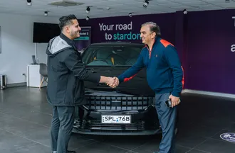 Two men shaking hands in a showroom, standing in front of a black Polestar with a visible license plate. Purple wall with text in the background.