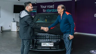 Two men shaking hands in a showroom, standing in front of a black Polestar with a visible license plate. Purple wall with text in the background.