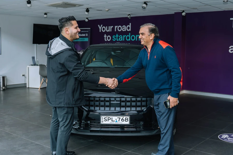 Two men shaking hands in a showroom, standing in front of a black Polestar with a visible license plate. Purple wall with text in the background.