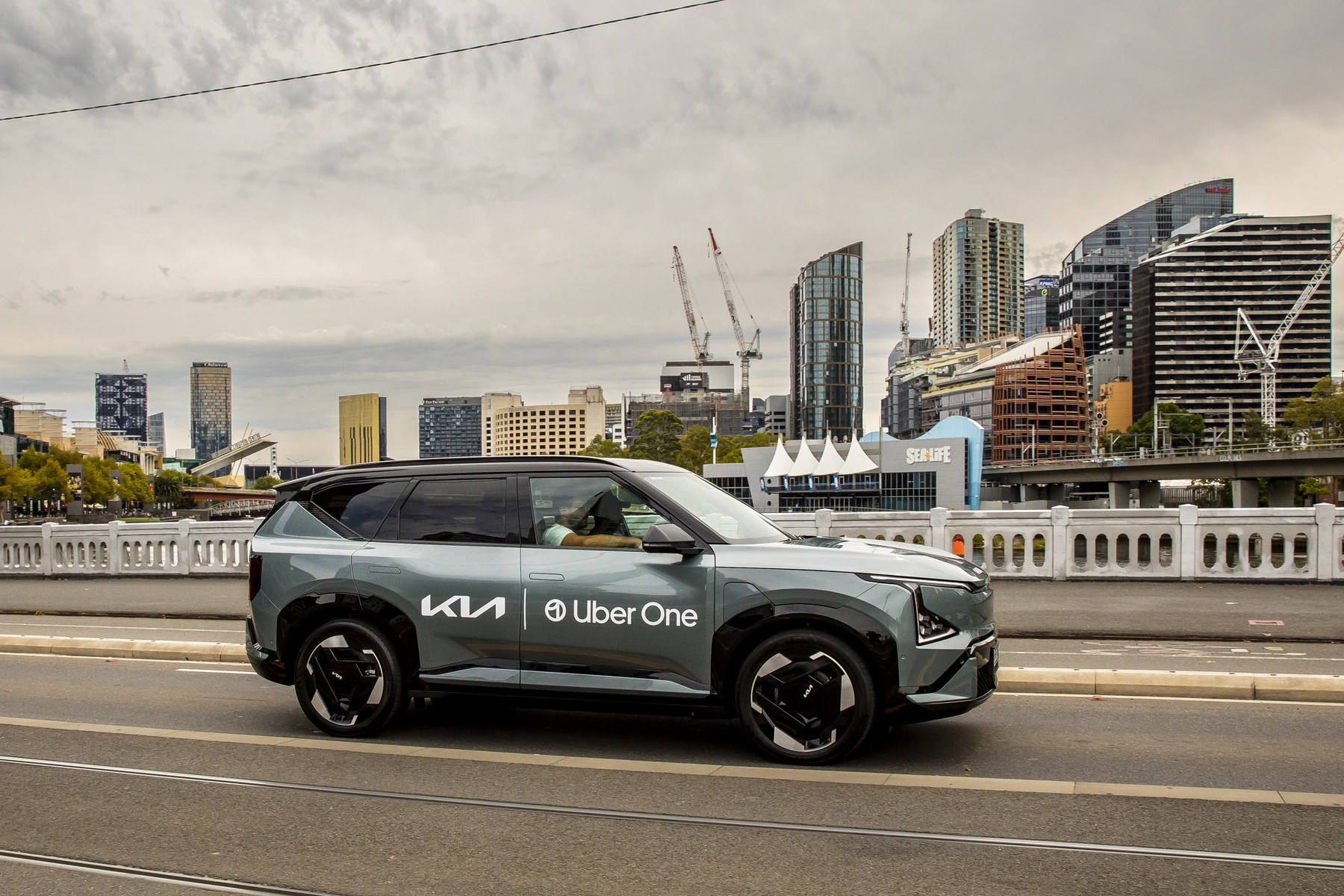 A Kia SUV with Uber One branding drives on a city street, with modern skyscrapers and cranes visible in the background.
