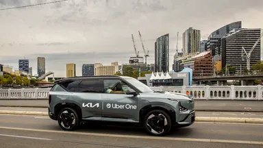 A Kia SUV with Uber One branding drives on a city street, with modern skyscrapers and cranes visible in the background.