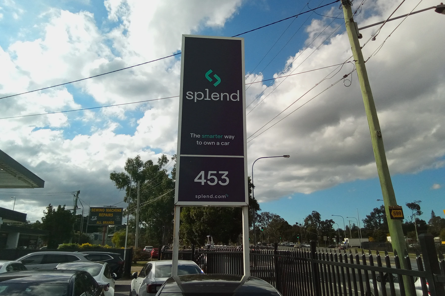 Sign in parking lot reads "splend: The smarter way to own a car" with website and number 453, against a partly cloudy sky.