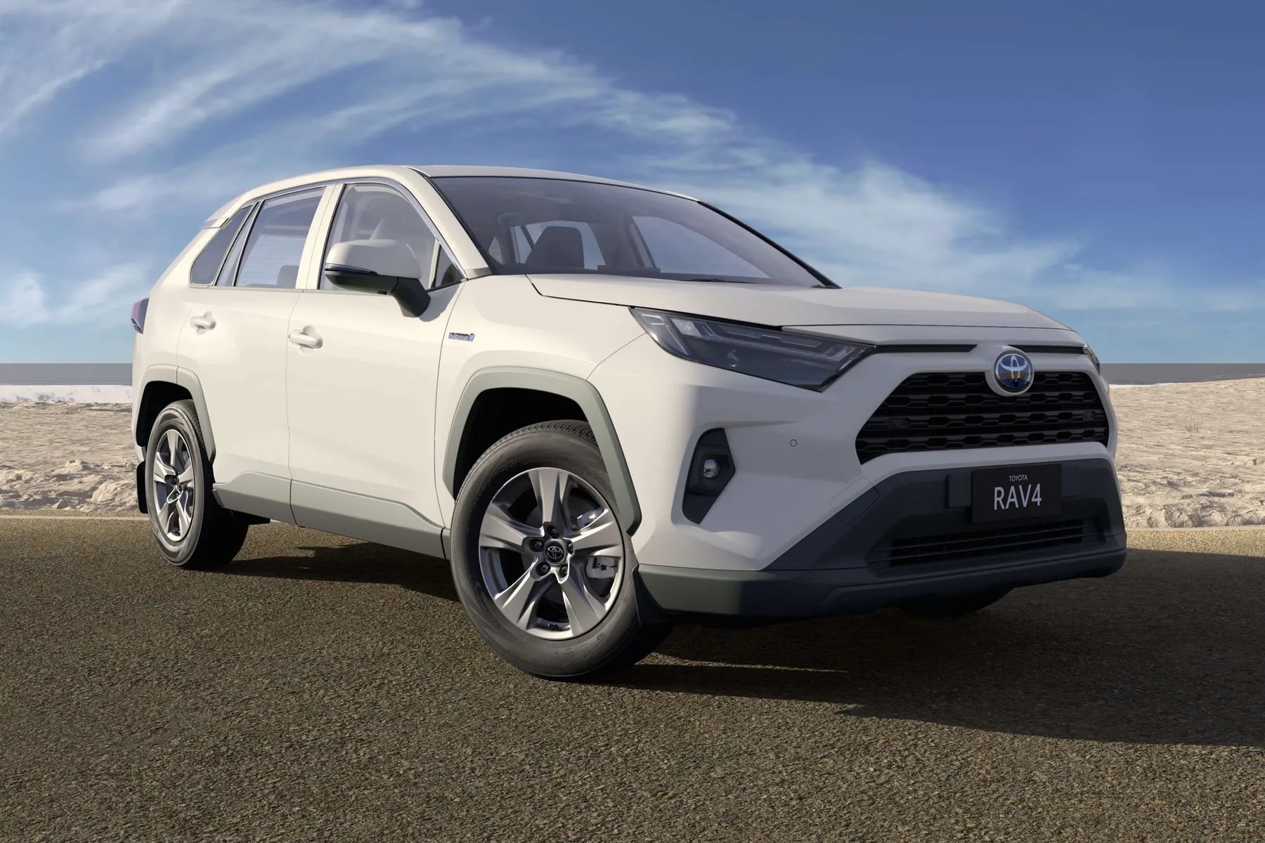White Toyota RAV4 parked on a paved road by a beach under a partly cloudy blue sky.