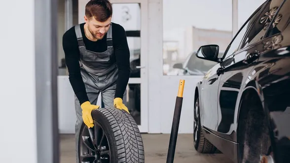 Mechanic in overalls and yellow gloves lifting a car tire inside a garage next to a black vehicle.
