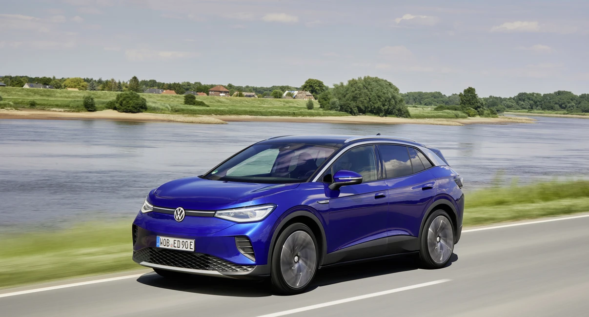 A blue Volkswagen ID.4 Pro SUV drives on a road beside a river, with green fields and trees in the background under a partly cloudy sky.