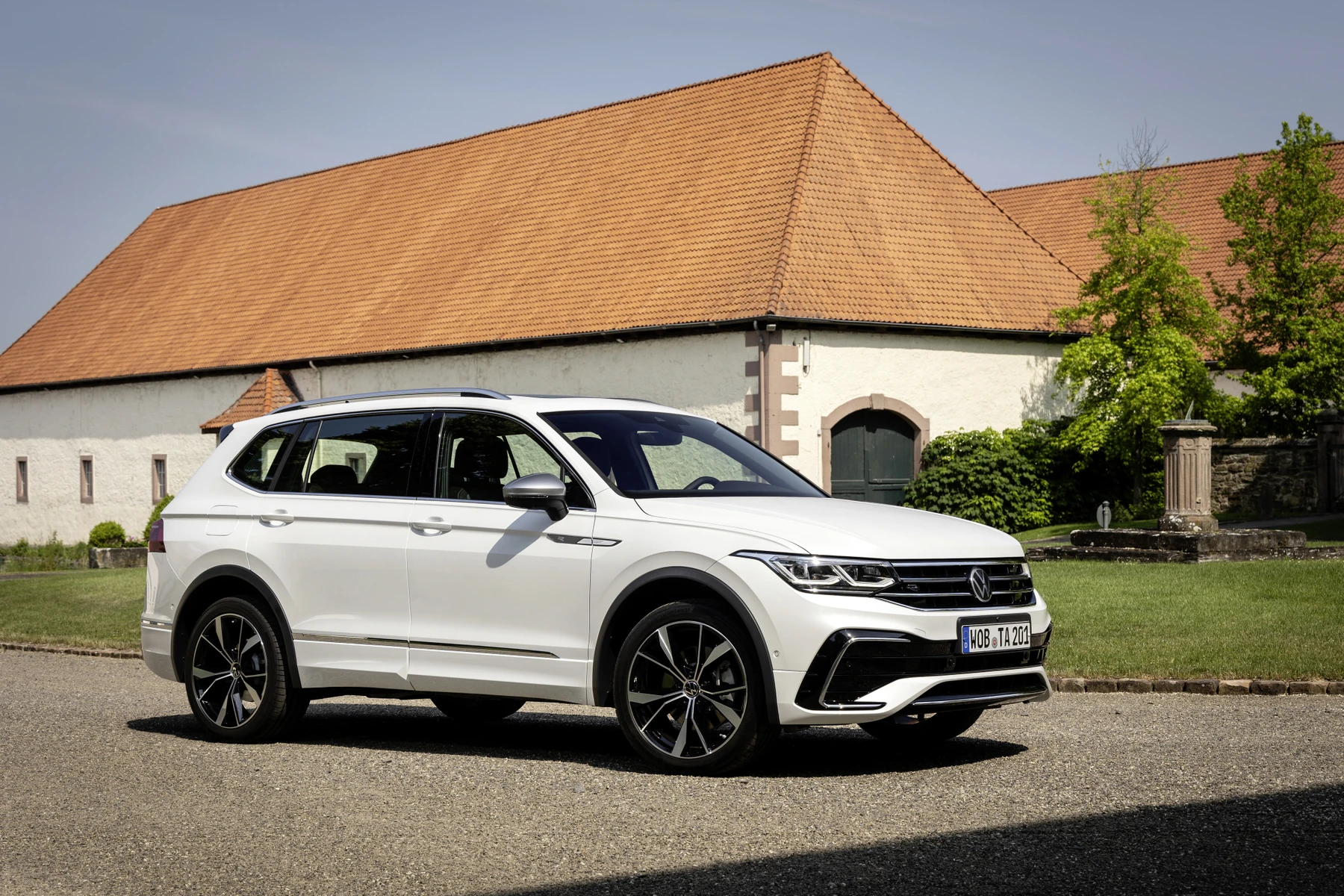White SUV parked on a paved area, with a rustic building and green trees in the background.