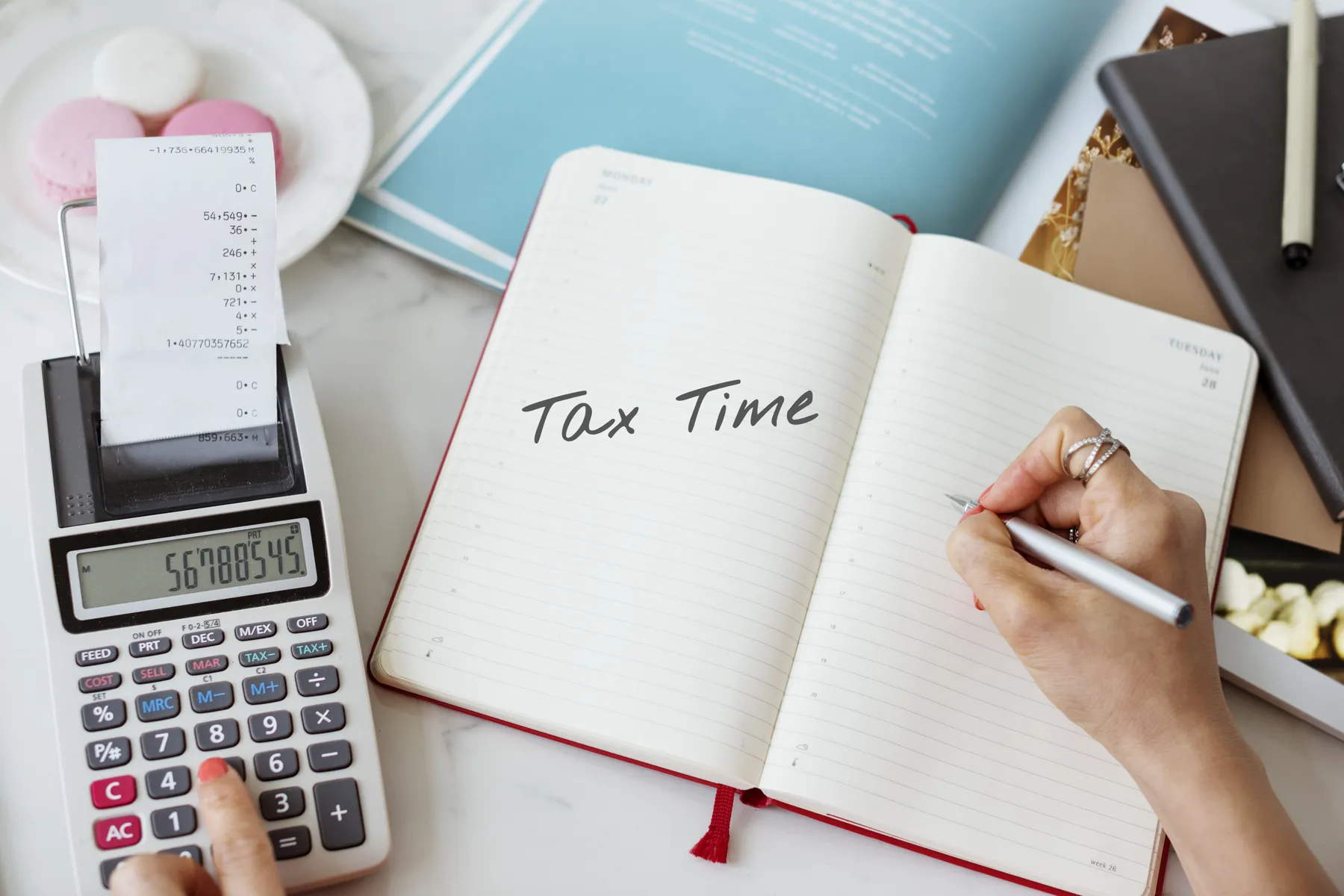 Open notebook with "Tax Time" written, calculator with receipt, and person writing on a desk with documents and macarons nearby.