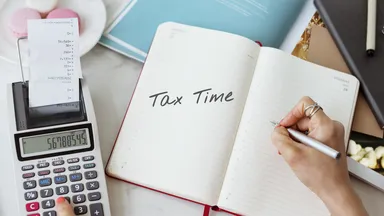 Open notebook with "Tax Time" written, calculator with receipt, and person writing on a desk with documents and macarons nearby.