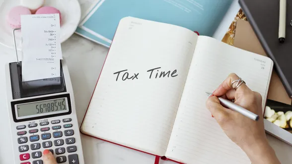 Open notebook with "Tax Time" written, calculator with receipt, and person writing on a desk with documents and macarons nearby.