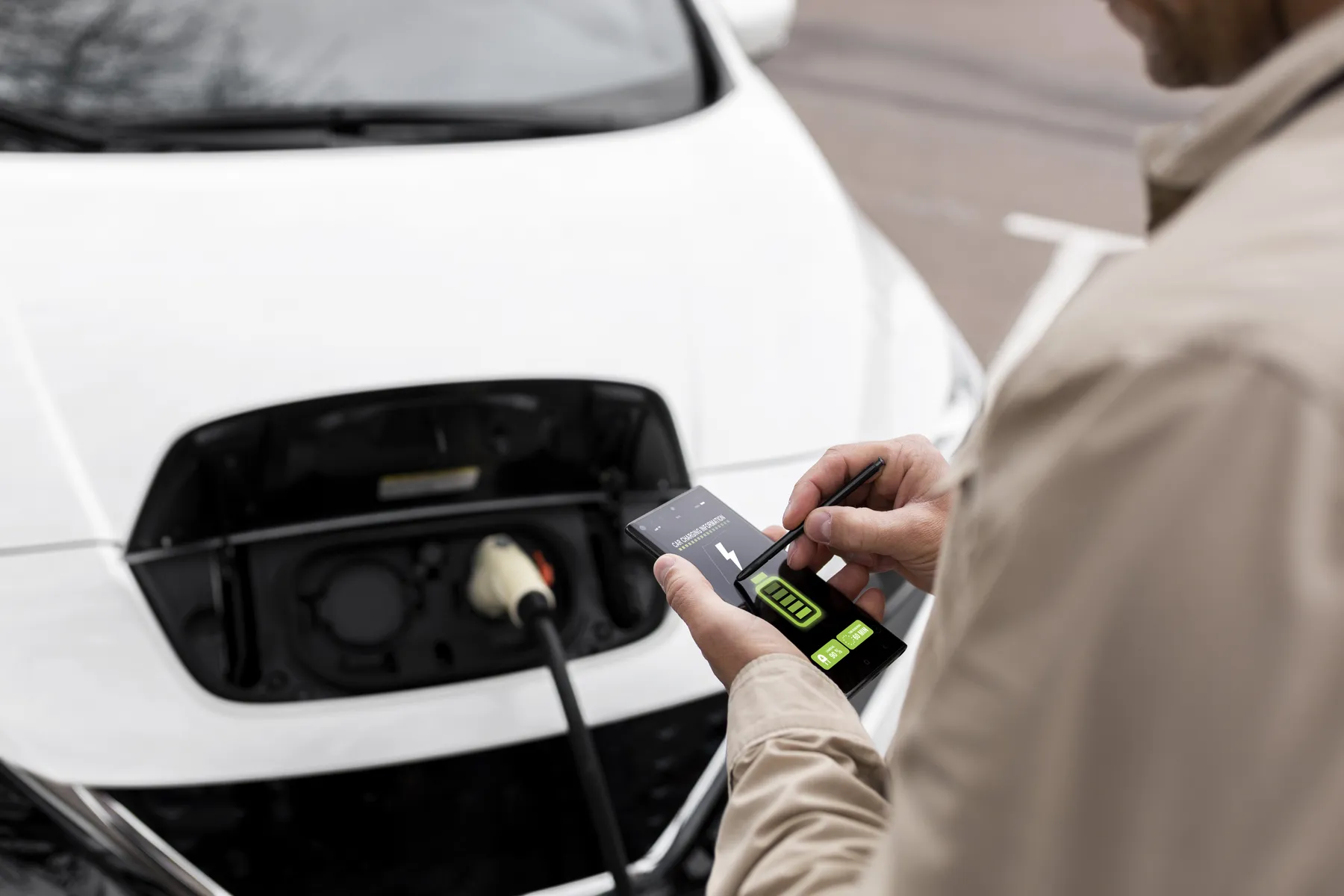 Person checks smartphone displaying battery/charging status while an electric car charges, cable plugged into the front charging port.