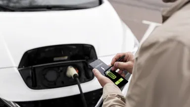 Person checks smartphone displaying battery/charging status while an electric car charges, cable plugged into the front charging port.