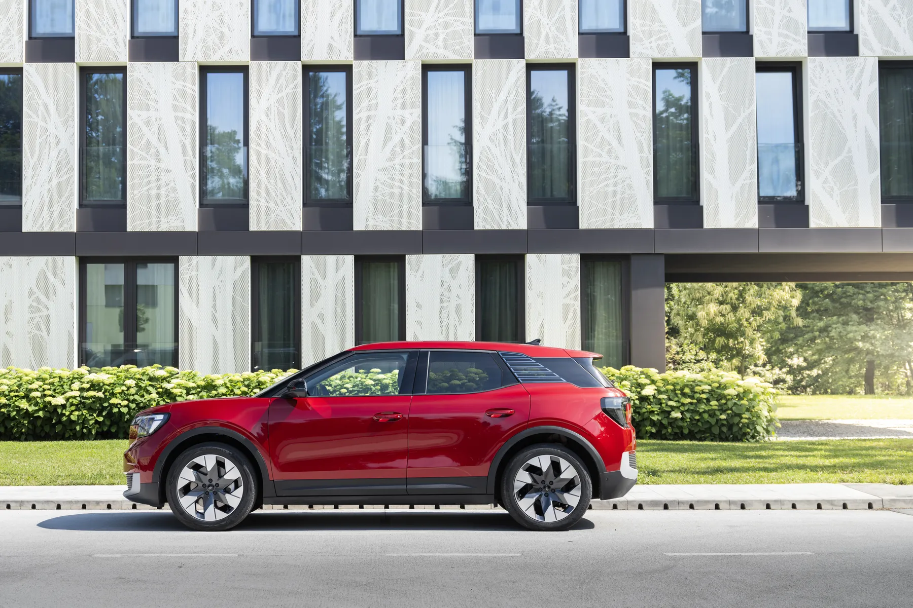 Red compact SUV parked on a street in front of a modern building with tall windows and a white tree-pattern facade.