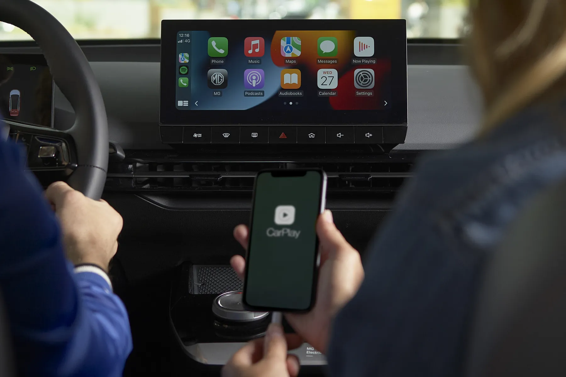 Passenger holds phone with CarPlay logo as driver steers; center dashboard shows Apple CarPlay app icons on the infotainment screen.