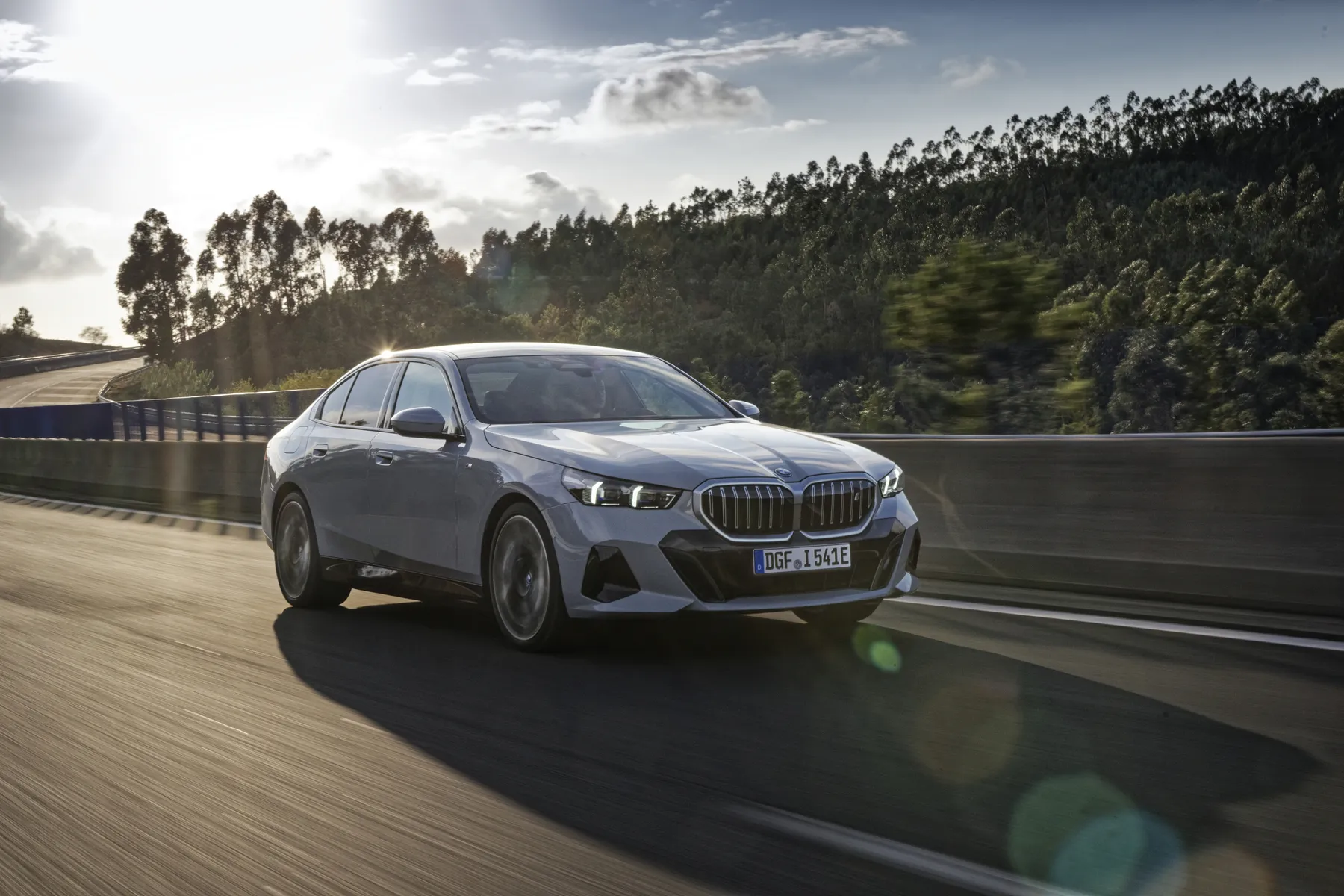 Silver sedan driving on a highway with trees in the background and the sun setting, casting a warm glow.