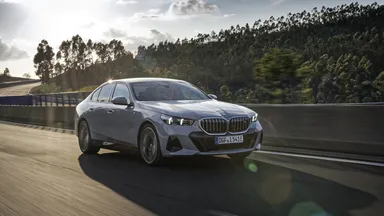 Silver sedan driving on a highway with trees in the background and the sun setting, casting a warm glow.