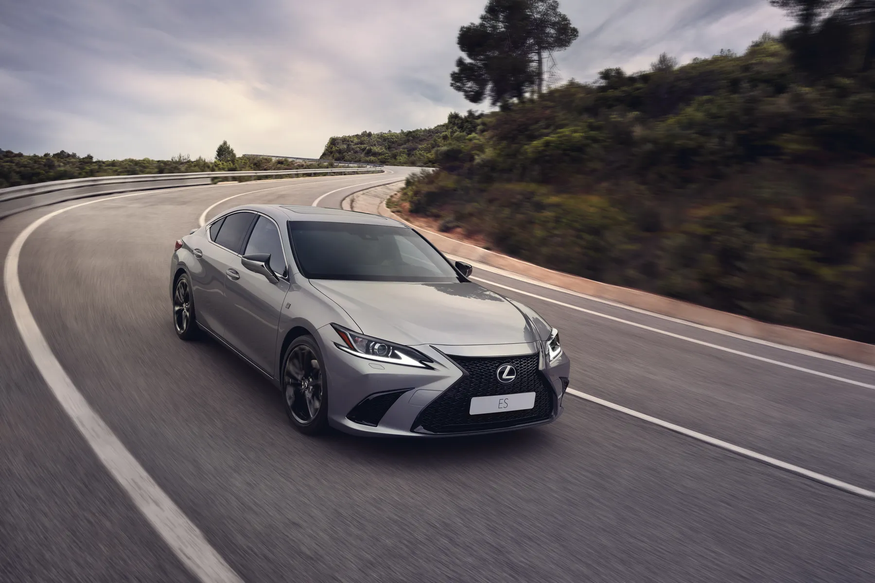 Silver Lexus ES sedan driving on a winding road with trees and hills in the background under a cloudy sky.