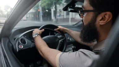 Man with a beard and glasses driving a car, viewed from the passenger side, with a street visible through the windshield.