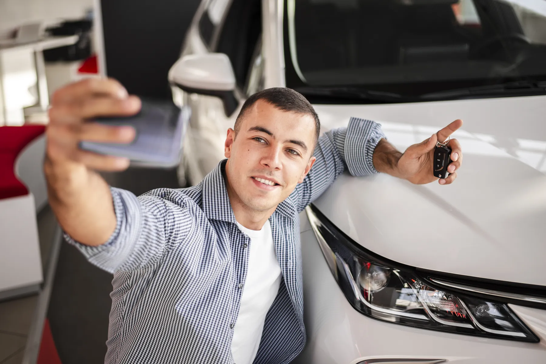 Man taking a selfie with a smartphone, smiling, while leaning on a white car and holding car keys.