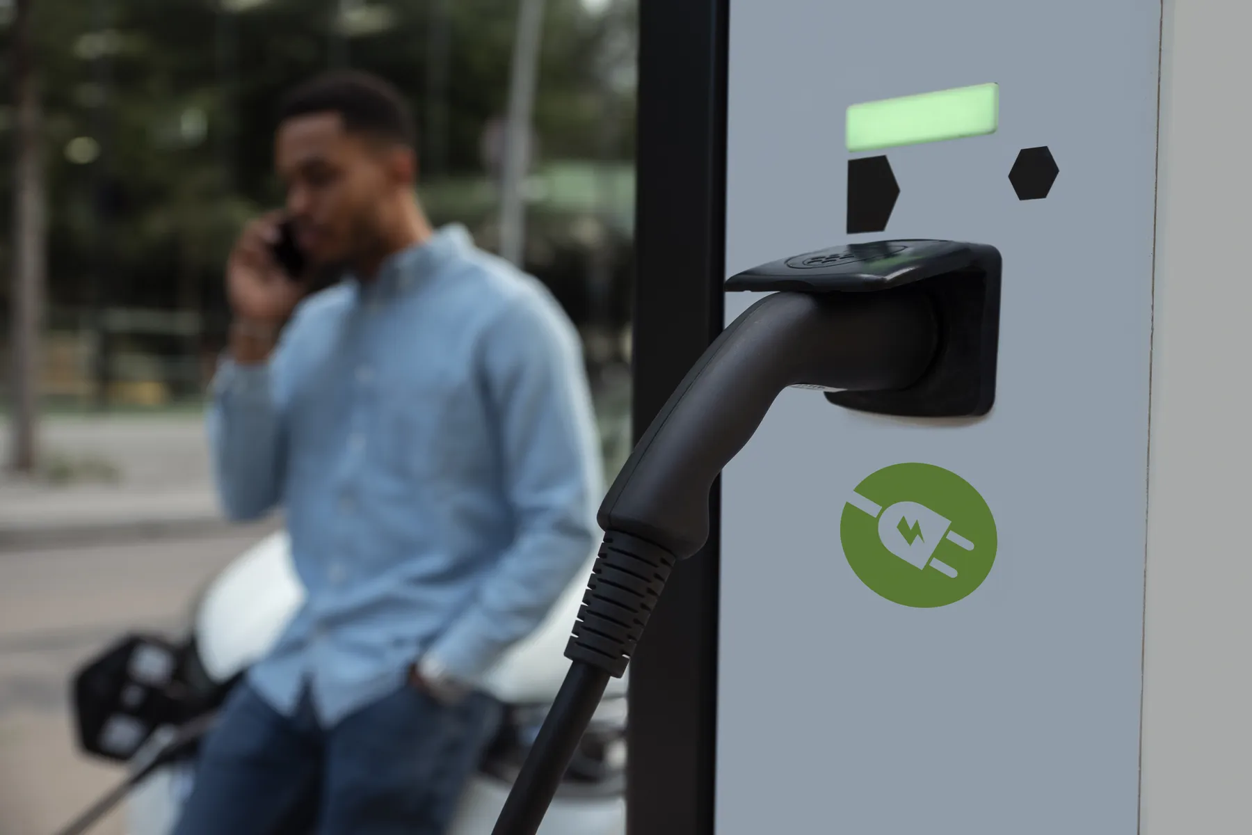 EV charging cable plugged into a station in foreground; blurred man on phone leaning against a parked car in the background.