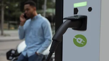 EV charging cable plugged into a station in foreground; blurred man on phone leaning against a parked car in the background.