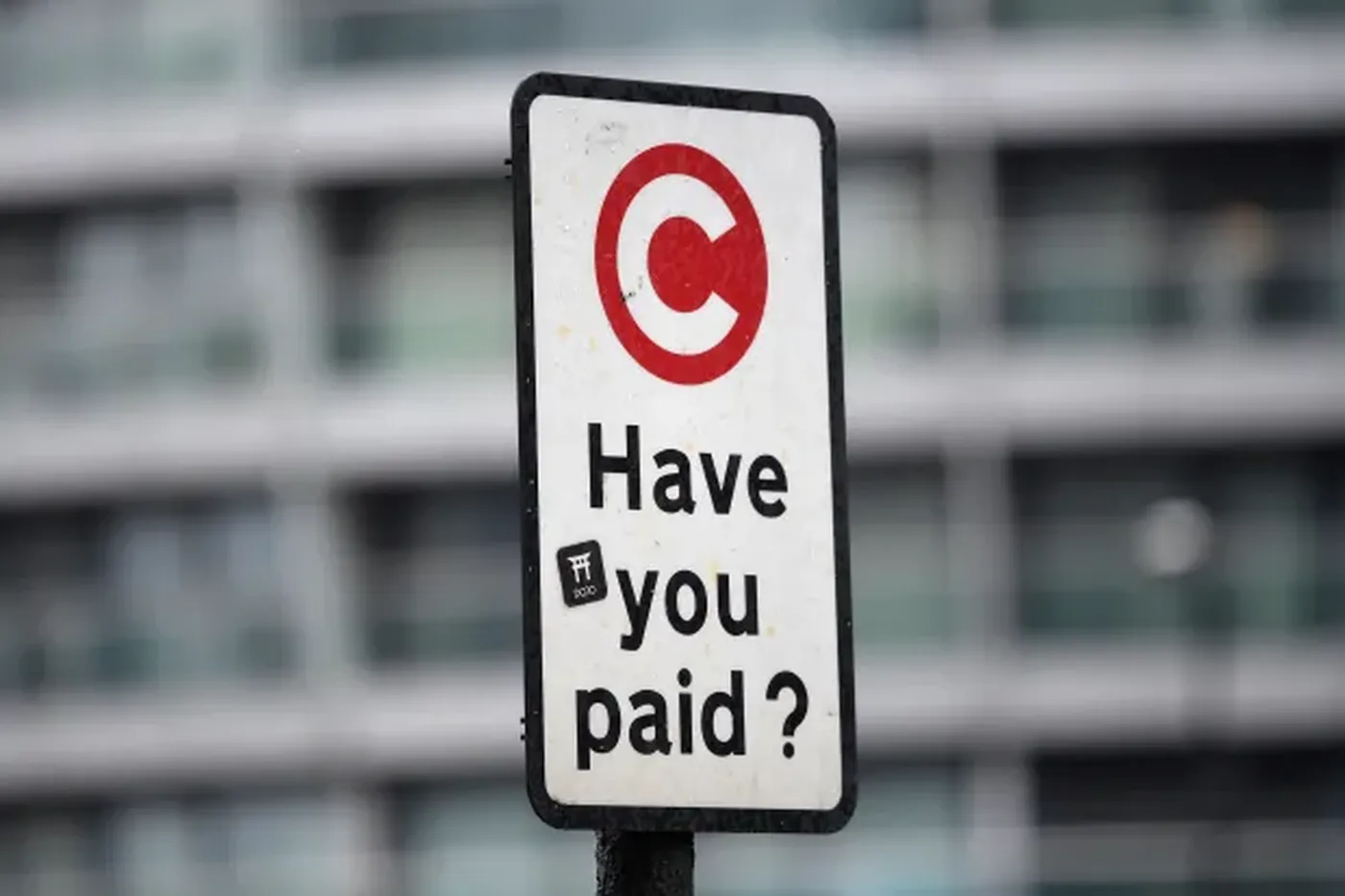 A sign with a red circle and "C" symbol reads "Have you paid?" against a blurred building background.