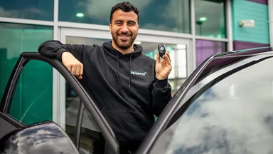 Smiling man leaning on an open car door, holding a car key fob in front of a glass dealership entrance.