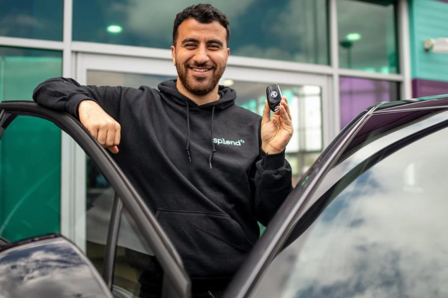 Smiling man leaning on an open car door, holding a car key fob in front of a glass dealership entrance.