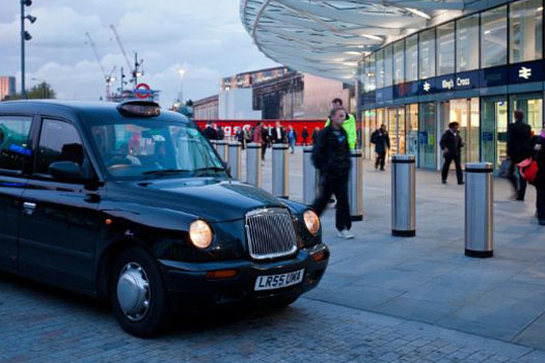 A black taxi is parked outside King's Cross station in London, with people walking nearby and a modern building in the background.