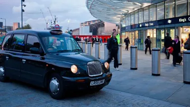 A black taxi is parked outside King's Cross station in London, with people walking nearby and a modern building in the background.