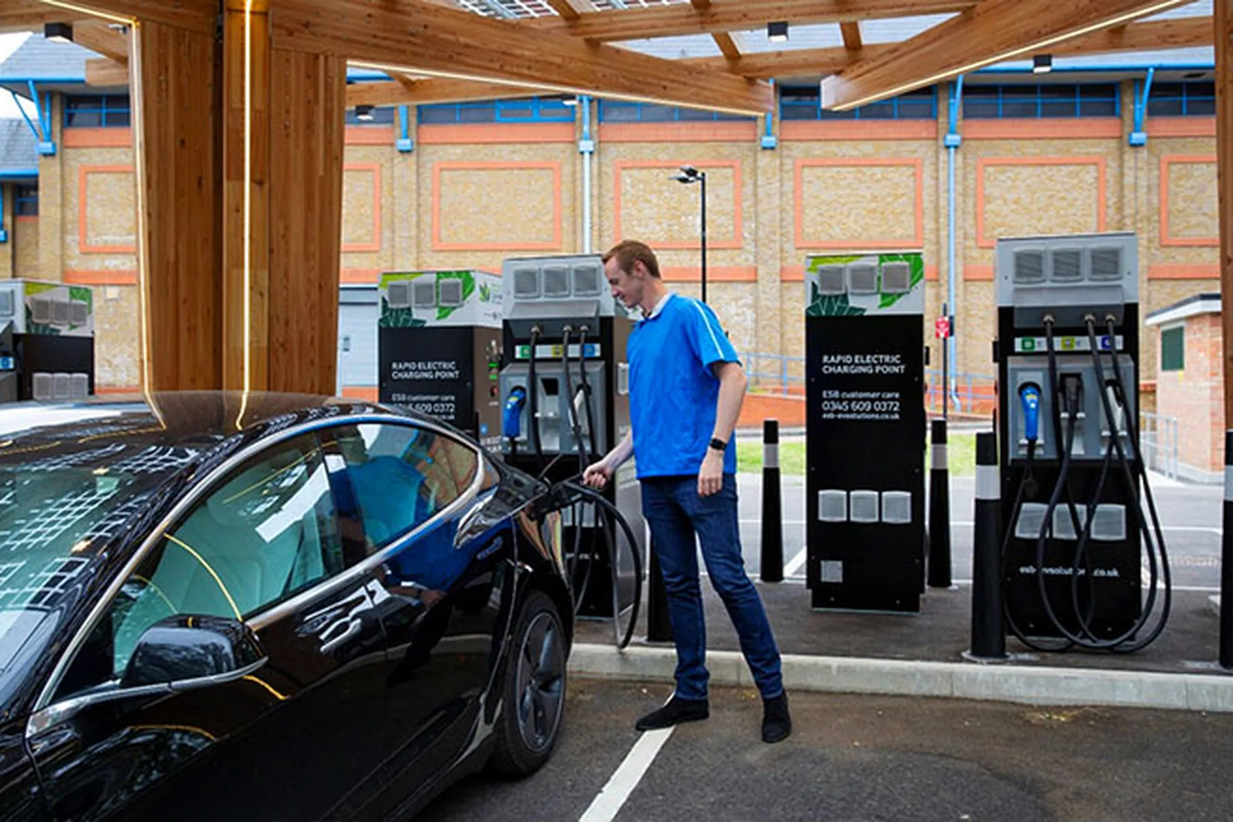Person in a blue shirt charging an electric car at a station with multiple charging units under a wooden canopy.