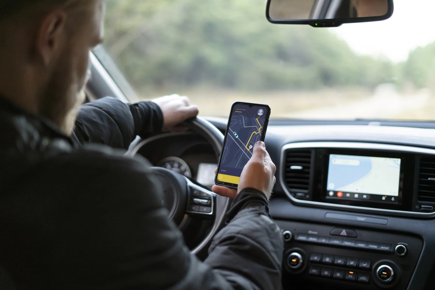 Driver holding a smartphone with a navigation map while driving, car dashboard and infotainment screen visible.