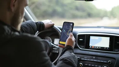 Driver holding a smartphone with a navigation map while driving, car dashboard and infotainment screen visible.