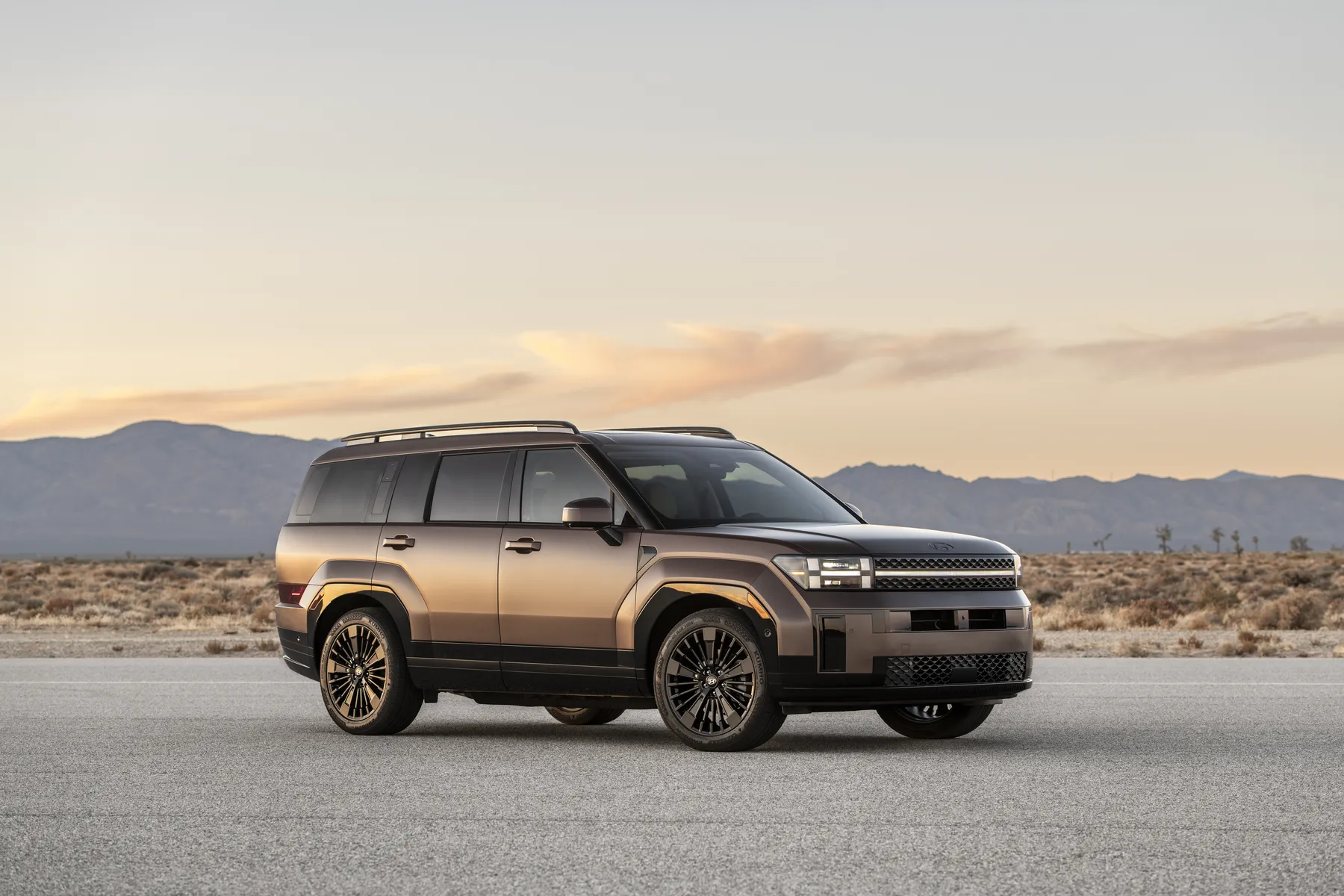 A modern SUV with a matte finish parked on an open road with a desert landscape and mountains in the background at sunset.