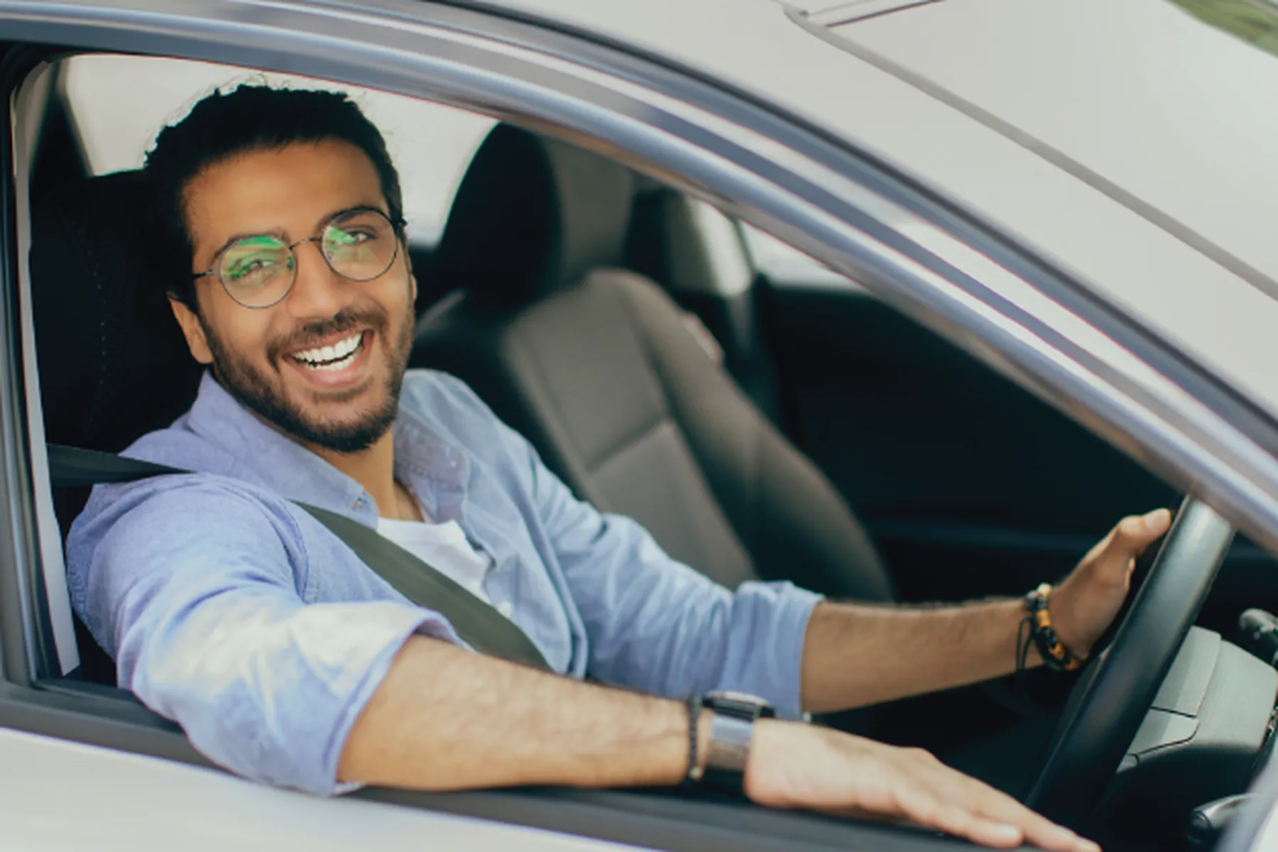 Smiling man with glasses and beard, wearing a light blue shirt and seatbelt, sitting in driver's seat with hand on the steering wheel.