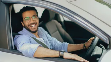 Smiling man with glasses and beard, wearing a light blue shirt and seatbelt, sitting in driver's seat with hand on the steering wheel.