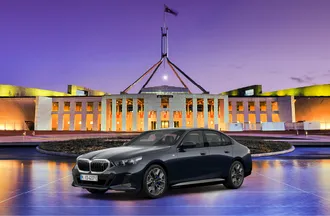 A sleek black BMW sedan is parked in front of a brightly lit, modern government building in Canberra at dusk.