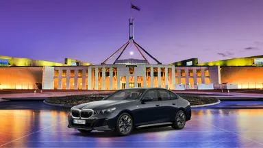 A sleek black BMW sedan is parked in front of a brightly lit, modern government building in Canberra at dusk.
