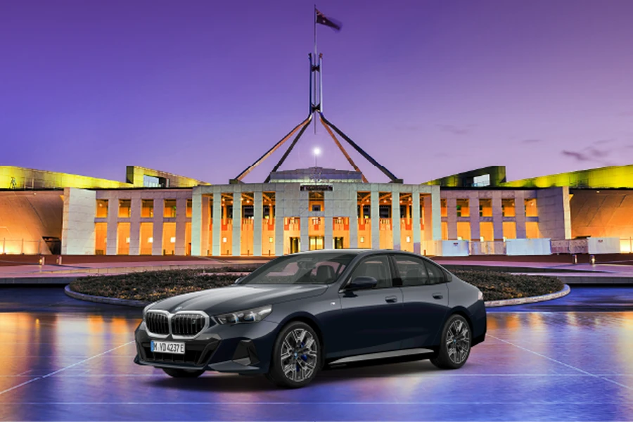 A sleek black BMW sedan is parked in front of a brightly lit, modern government building in Canberra at dusk.