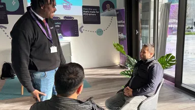 Three men in a modern office: one standing and speaking to two seated colleagues by a window, with plants and a coffee cup nearby.