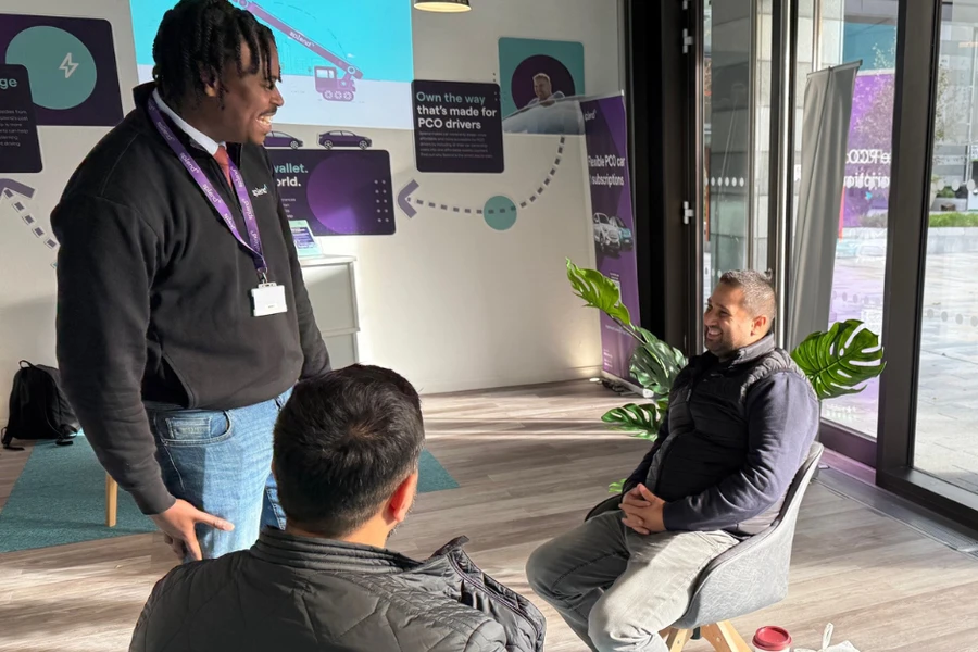 Three men in a modern office: one standing and speaking to two seated colleagues by a window, with plants and a coffee cup nearby.