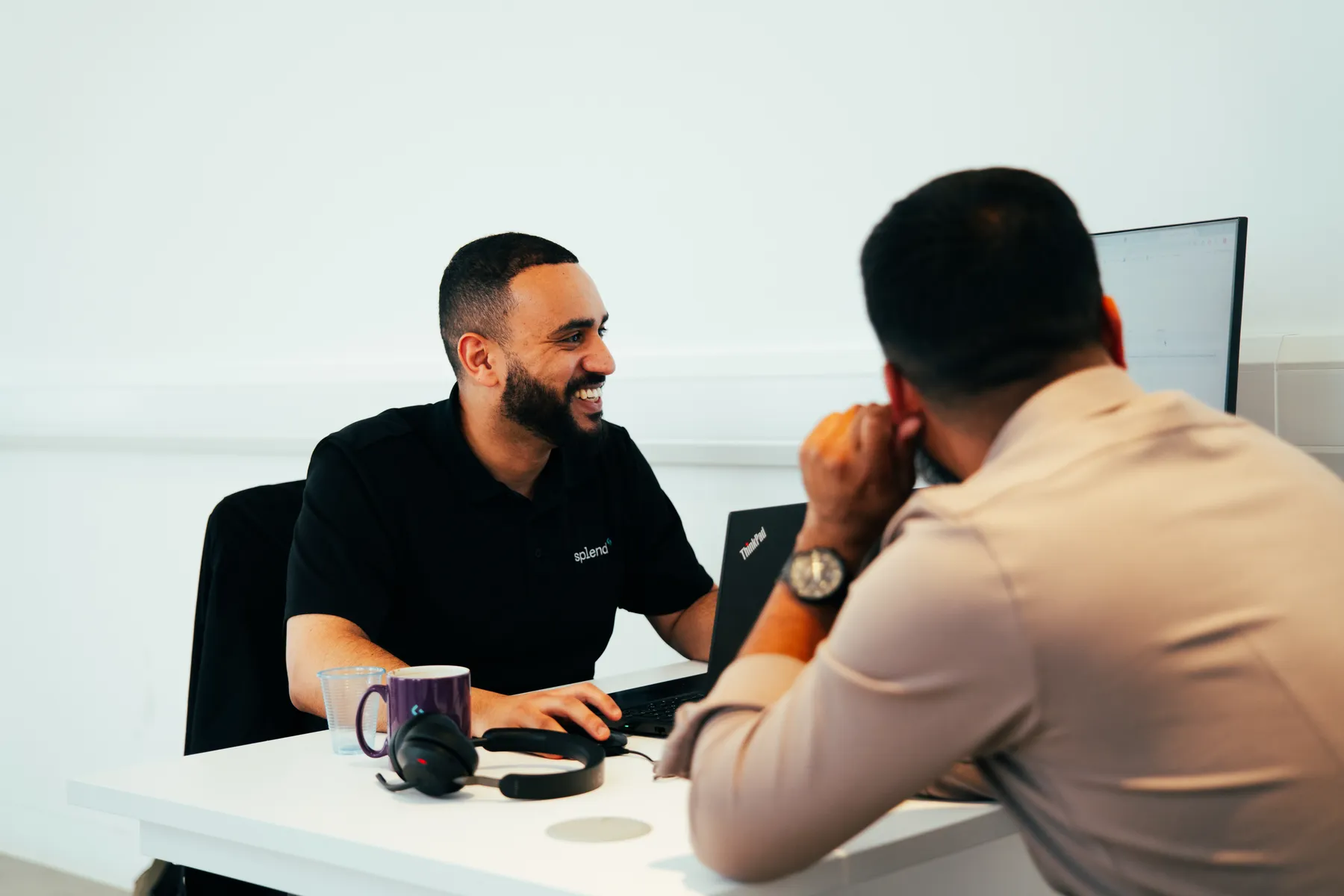 Two men are seated at a desk, engaged in conversation. One is smiling, using a laptop. A mug and headphones rest nearby.