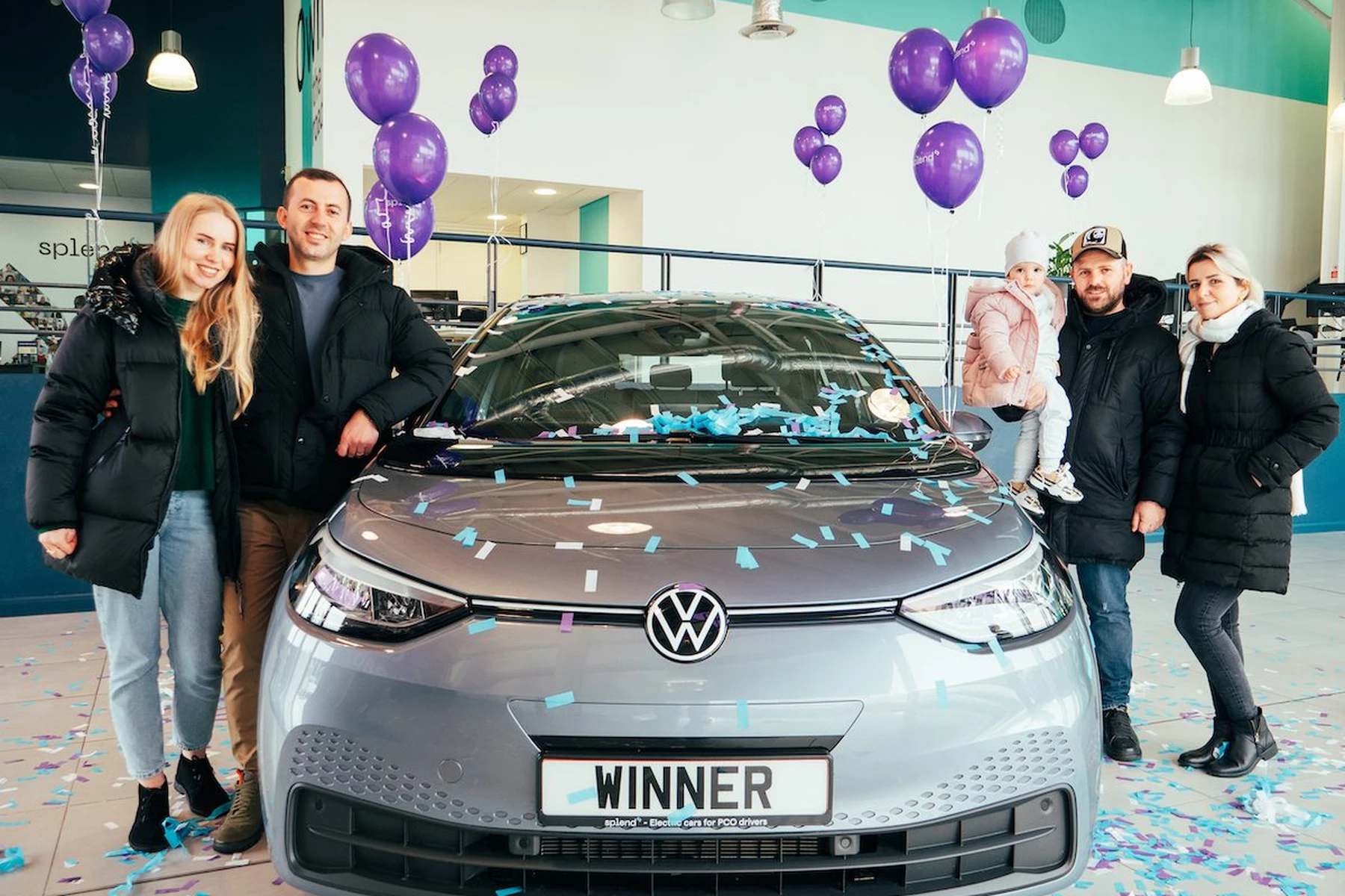 A group of people stands beside a Volkswagen car with a "WINNER" plate, surrounded by purple balloons and confetti in a showroom.