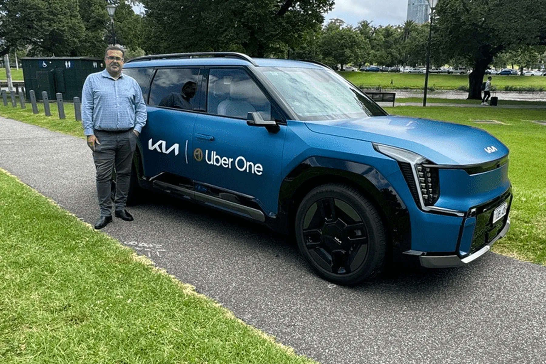 Man standing next to a blue Kia Uber One car parked on a road, with trees and grass in the background.
