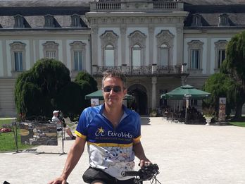 Christian Weinberger in cycling gear poses with a bicycle in front of a grand historic building on a sunny day.