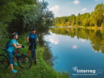 Zwei Radfahrer:innen machen am grünen Flussufer Pause; stilles Wasser spiegelt Auwald und blauen Himmel.